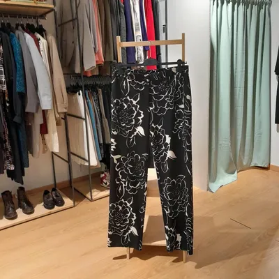 Pantalone Camomilla, nero con fiori bianchi, a gamba ampia, ha un bottone nero con riportata la firma ed una cerniera ch